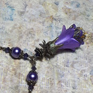 Vintage Style Deadly nightshade inspired necklace that has a purple flower drop with topaz yellow crystals and antique floral bead caps. It has a bronze chain that has purple pearls and crystals spaced along it. It has a whimsigoth style to it.