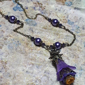 Vintage Style Deadly nightshade inspired necklace that has a purple flower drop with topaz yellow crystals and antique floral bead caps. It has a bronze chain that has purple pearls and crystals spaced along it. It has a whimsigoth style to it.