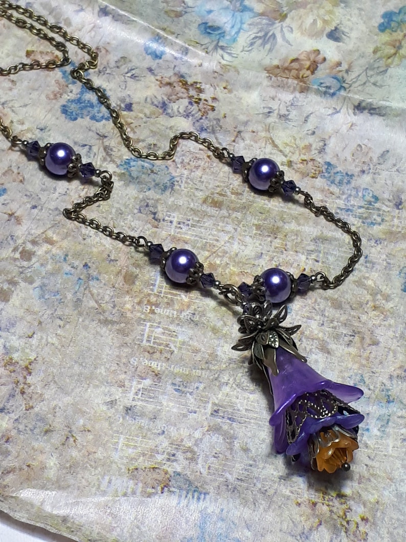 Vintage Style Deadly nightshade inspired necklace that has a purple flower drop with topaz yellow crystals and antique floral bead caps. It has a bronze chain that has purple pearls and crystals spaced along it. It has a whimsigoth style to it.
