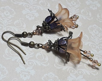 May include: A pair of dangle earrings featuring a delicate flower design. The flowers are made of a translucent peach-colored material and have a bronze-colored metal base. The earrings are accented with small, clear crystals and have a bronze-colored hook.