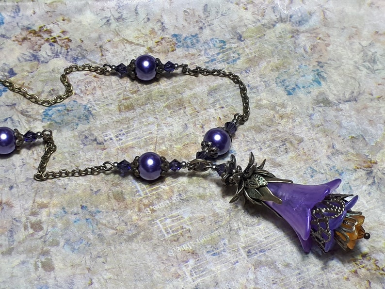 Vintage Style Deadly nightshade inspired necklace that has a purple flower drop with topaz yellow crystals and antique floral bead caps. It has a bronze chain that has purple pearls and crystals spaced along it. It has a whimsigoth style to it.