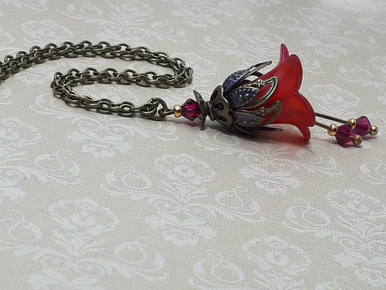 A red vintage style Lucite flower pendant necklace with a bronze filigree design and Siam red crystal beads that hang and sway from under the flowers. The necklace has a bronze tone chain. Fairycore style jewelry gift. Whimsical Necklace.