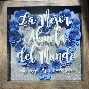 May include: A framed shadow box with a heart made of blue paper flowers. The text "La Mejor Abuela del Mundo" is written in white on a silver glitter background. The names Abi, Angel, Amy, Isaiah, and Yesenia are written below the heart.
