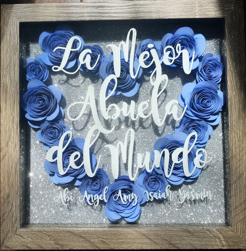May include: A framed shadow box with a heart made of blue paper flowers. The text "La Mejor Abuela del Mundo" is written in white on a silver glitter background. The names Abi, Angel, Amy, Isaiah, and Yesenia are written below the heart.