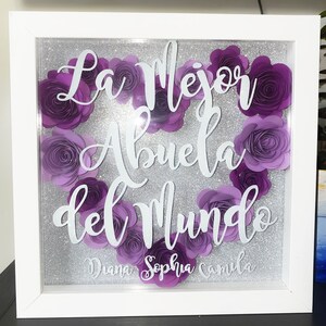 May include: A white frame with a silver glitter background and purple paper flowers. The text "La Mejor Abuela del Mundo" is written in white on the frame. The names "Diana, Sophia, Camila" are written in white at the bottom of the frame.
