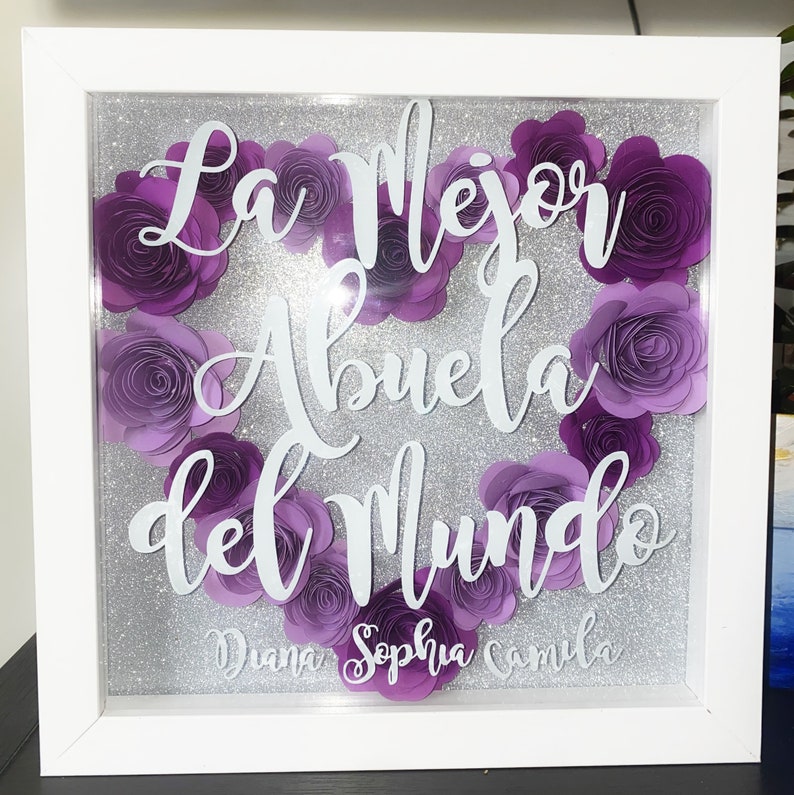 May include: A white frame with a silver glitter background and purple paper flowers. The text "La Mejor Abuela del Mundo" is written in white on the frame. The names "Diana, Sophia, Camila" are written in white at the bottom of the frame.
