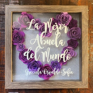 May include: A framed shadow box with a heart-shaped arrangement of purple paper flowers. The text "La Mejor Abuela del Mundo" is displayed in white lettering. The names "Graciela, Osvaldo, Sofia" are written below the heart.