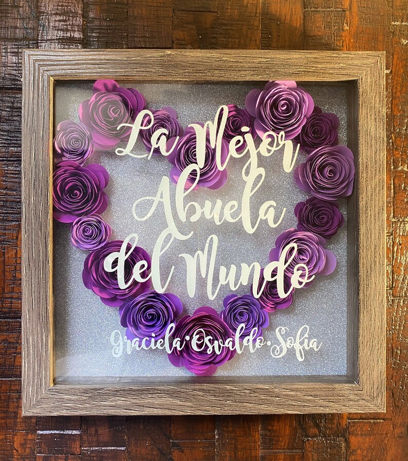 May include: A framed shadow box with a heart-shaped arrangement of purple paper flowers. The text "La Mejor Abuela del Mundo" is displayed in white lettering. The names "Graciela, Osvaldo, Sofia" are written below the heart.