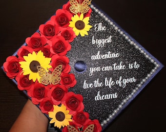Può includere: Un cappello di laurea blu navy decorato con rose di carta rossa, girasoli gialli e farfalle dorate. Il cappello ha uno sfondo di glitter bianco e una citazione bianca che recita "La più grande avventura che puoi intraprendere è vivere la vita dei tuoi sogni."