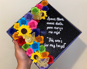 May include: A navy blue graduation cap decorated with colorful paper flowers, butterflies, and sunflowers. The cap has the text "Aveces lloré, aveces dud'e pero nunca me razé" and "This one's for my boys!" written in white.