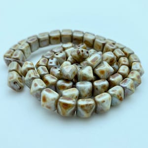 Puede incluir: Una hebra de cuentas de vidrio blancas y marrones con un acabado moteado. Las cuentas tienen forma de pequeñas pirámides irregulares.