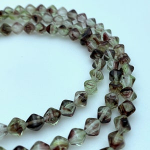 Puede incluir: Hebras de cuentas facetadas en tonos verdes, marrones y transparentes. Las cuentas tienen forma cuadrada con bordes redondeados y un aspecto veteado. Las cuentas están ensartadas, creando un collar o pulsera.