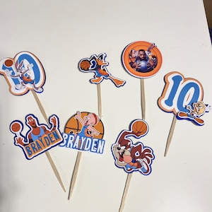 May include: Set of 10 cupcake toppers featuring characters from Space Jam 2. The toppers include Bugs Bunny, Daffy Duck, Lola Bunny, and the Tune Squad. The toppers are printed on cardstock and have wooden sticks.