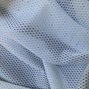 Puede incluir: Tela de malla blanca con un pequeño patrón repetitivo de agujeros ovalados. La tela está doblada y drapeada, mostrando la textura del material.