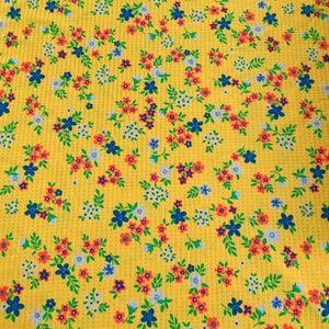 May include: Yellow fabric with a repeating floral pattern. The design features small blue, orange, and white flowers with green leaves. The fabric has a textured, waffle-like appearance.