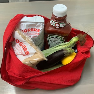 May include: A red reusable shopping bag filled with groceries, including two bags of sugar, a bottle of Heinz tomato ketchup, corn on the cob, an eggplant, and a lemon.