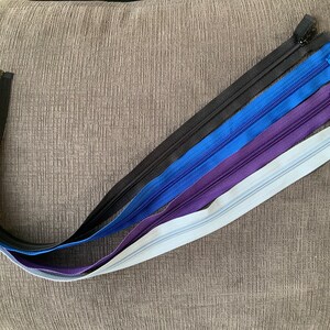 Puede incluir: Un conjunto de seis cremalleras en varios tonos de azul, morado y negro. Las cremalleras están dispuestas en forma de arcoíris, con la cremallera negra en la parte superior y la cremallera azul claro en la parte inferior.