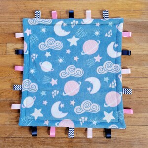 May include: A square baby blanket with a blue background and a space-themed pattern of planets, stars, clouds, and moons. The blanket has a border of various colored ribbons, including pink, navy, and black and white zig-zag ribbons.
