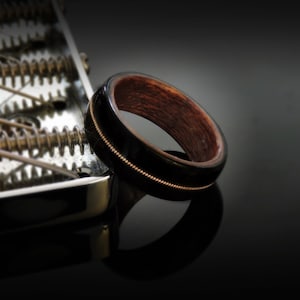 May include: A wooden ring with a black inlay and a thin gold band. The ring is made of dark wood and has a polished finish.