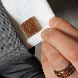May include: A pair of wooden cufflinks with a brown wood grain pattern. The cufflinks are square shaped and have a subtle sheen.