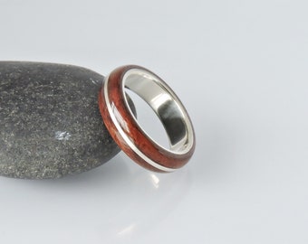 Op de afbeelding: Een houten ring met een zilveren band. Het hout is donker roodbruin met een lichtere bruine streep die rond de ring loopt. De ring ligt op een grijze steen.