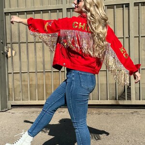 Pode incluir: Casaco vermelho com franjas douradas e a palavra "CHIEFS" nas costas. O casaco tem estrelas douradas e detalhes de penas. A pessoa est&aacute; usando jeans azuis e botas brancas com salto bloco.