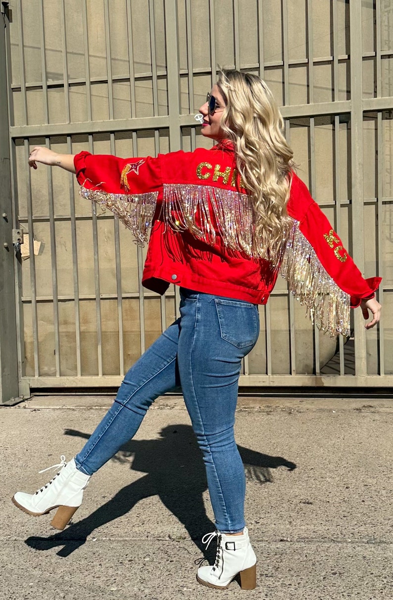 Pode incluir: Casaco vermelho com franjas douradas e a palavra "CHIEFS" nas costas. O casaco tem estrelas douradas e detalhes de penas. A pessoa est&aacute; usando jeans azuis e botas brancas com salto bloco.