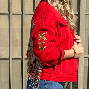 Pode incluir: Casaco de ganga vermelho com letras douradas "KC" de lantejoulas na manga. A parte de tr&aacute;s do casaco apresenta longas franjas prateadas. O casaco tem gola cl&aacute;ssica, bot&otilde;es e bolsos frontais. A modelo usa cal&ccedil;as de ganga azuis.