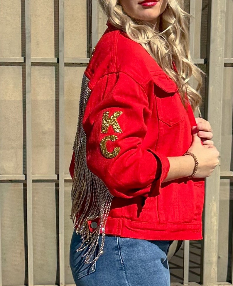 Pode incluir: Casaco de ganga vermelho com letras douradas "KC" de lantejoulas na manga. A parte de tr&aacute;s do casaco apresenta longas franjas prateadas. O casaco tem gola cl&aacute;ssica, bot&otilde;es e bolsos frontais. A modelo usa cal&ccedil;as de ganga azuis.