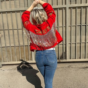 Pode incluir: Casaco de ganga vermelho com detalhes de estrelas e franjas douradas. A palavra "CHEERS" est&aacute; escrita em letras douradas nas costas. O casaco &eacute; combinado com jeans azuis e botas brancas.
