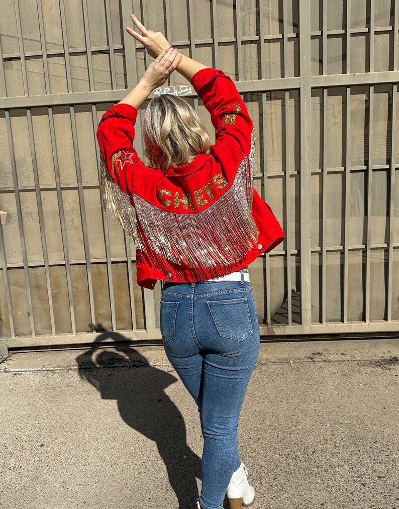Pode incluir: Casaco de ganga vermelho com detalhes de estrelas e franjas douradas. A palavra "CHEERS" est&aacute; escrita em letras douradas nas costas. O casaco &eacute; combinado com jeans azuis e botas brancas.