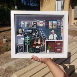 May include: A shadow box diorama with a white frame, depicting an office scene with a pilot at a desk. The background shows an airport runway, a clock, and travel destination signs. Miniature luggage and a plant are also present.