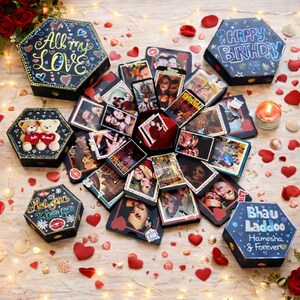 May include: A collection of hexagonal gift boxes, some open to reveal photos, scattered on a wooden surface. The boxes are black with decorative text like "All my LOVE" and "Happy Birthday." Red heart-shaped confetti and a lit candle add to the festive scene.