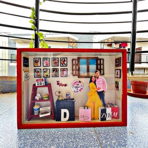 May include: A red framed shadow box with miniature items, including photos, a ladder shelf with food, and a couple figurine. The box has a window and a small table with the letter "D". Shopping bags are also included.