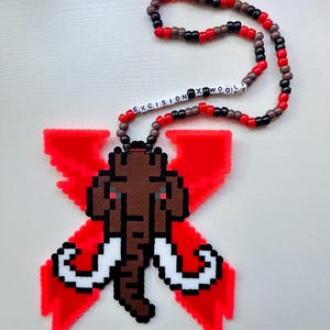 May include: A necklace featuring a pendant made of red, brown, white, and black plastic beads arranged in a pixelated mammoth shape over a red X symbol. The necklace strand consists of red, black, and brown beads with white letter beads spelling EXCISION X WOOLI.