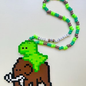 Puede incluir: Un collar de cuentas con un colgante pixelado de un dinosaurio verde montado sobre un mamut marrón. El collar está hecho de cuentas de plástico verde lima y marrón, con cuentas de letras blancas que deletrean WOOLI X EXCISION.