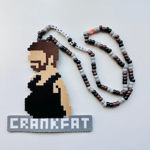 May include: A pixelated pendant necklace featuring a figure with a beard and the text "CRANKFAT". The necklace is made of black, brown, gray, and white beads. The pendant is black, brown, and tan.