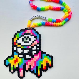 May include: A colorful kandi necklace with a pixelated pendant. The pendant features a rainbow-colored design with a single eye and the word "SUBTRONICS" spelled out in white beads. The necklace is made of rainbow-colored beads.