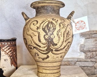 Pode incluir: Ânfora de cerâmica bege com um desenho de polvo castanho escuro. O vaso tem duas alças e uma boca larga. Uma etiqueta com um desenho floral pende de uma alça. O vaso é exibido em uma superfície de pedra.