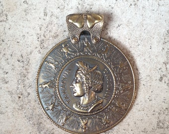 Pendant in burnished brass with the face of Tanaquilia in the center and a group of dancers and musicians