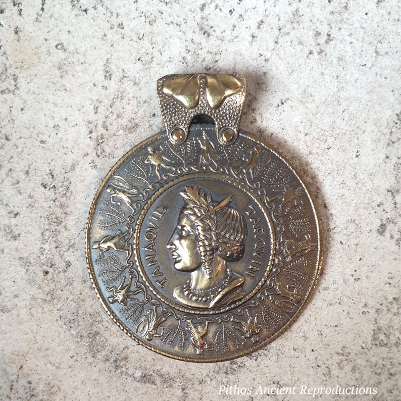 Pendant in burnished brass with the face of Tanaquilia in the center and a group of dancers and musicians image 1