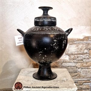 Peut inclure: Un vase Pithos en céramique noire avec un couvercle, des anses et une base en piédestal. Le vase présente des accents blancs et est étiqueté "Pithos Ancient Reproductions". Le vase est exposé sur une surface en pierre.