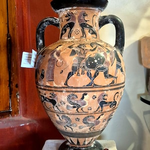Puede incluir: Una ánfora de cerámica color terracota con ilustraciones de estilo de figuras negras que representan figuras mitológicas, leones y aves. El recipiente tiene dos asas negras, un borde ancho y una base, con bandas decorativas alrededor del cuerpo.