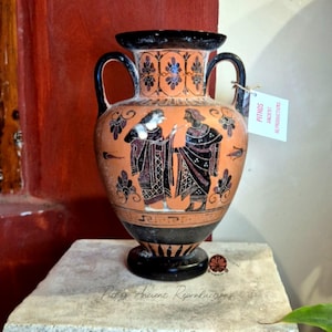 Puede incluir: Un jarrón ánfora de cerámica color terracota con figuras pintadas en negro con vestimenta tradicional y patrones geométricos complejos. El jarrón tiene dos asas laterales y una etiqueta que dice PITHOS ANCIENT REPRODUCTIONS.