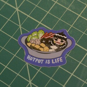 May include: A die-cut sticker featuring a cartoon character with dark hair sitting in a gray hotpot filled with green lettuce, sliced lotus root, brown mushrooms, tofu cubes, and red-striped meat. The text HOTPOT IS LIFE is printed in white.