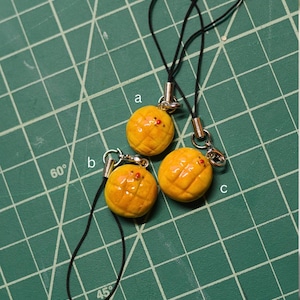 May include: Three miniature, yellow, melon-shaped charms with black cords and silver-colored metal clasps. The charms have a textured surface and small red accents. The background is a green cutting mat.