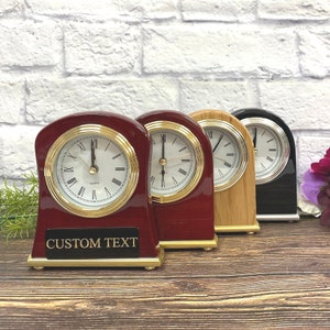 May include: Four tabletop clocks with different colored bases. The clocks have gold trim and Roman numeral faces. The clock on the left has a red base and a black plaque with the text "CUSTOM TEXT".