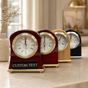 May include: A row of four arched mantel clocks with white faces, black Roman numerals, and gold-tone bezels. The first clock is dark mahogany with a black plaque reading CUSTOM TEXT. The others are cherry, light oak, and black, all on gold feet.