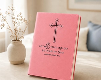 May include: A pink hardcover journal featuring a black graphic of a cross centered above the text, "LET ALL THAT YOU DO BE DONE IN love CORINTHIANS 16:14." The journal is positioned upright, displaying its front cover with a smooth, matte texture.