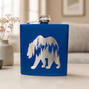 May include: A royal blue metal flask featuring a silver-toned engraved silhouette of a bear. The bear's body is filled with a detailed landscape design showing a dense forest of pine trees and mountain peaks. It has a hinged silver screw cap.
